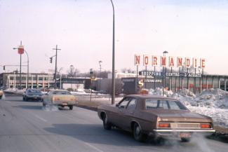 Photo couleur montrant un taxi et d'autres véhicules circulant dans une rue assez large. En arrière-plan, une enseigne annonce « NORMANDIE. SHOPPING CENTRE D’ACHATS » et on peut voir un supermarché Steinberg.