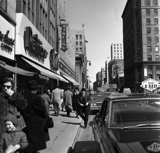 Photo noir et blanc de la rue Sainte-Catherine montrant les magasins sur la gauche, des passants sur le trottoir et des voitures et camions dans la rue.