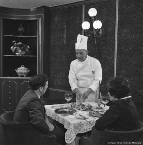 Photographie en noir et blanc représentant un couple à table, de dos, devant lequel se tient un chef chinois qui s’apprête à leur servir un plat. 