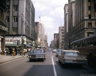 Photo couleur montrant la rue Sainte-Catherine, au centre-ville de Montréal, avec des voitures et des autobus qui circulent dans la rue, une foule au coin de la rue Peel, et les édifices de chaque côté.  