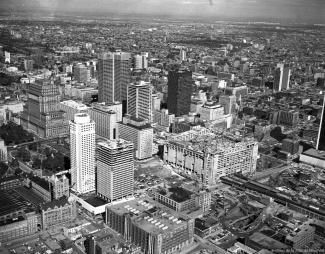 Photographie en noir et blanc du panorama montréalais d’un point de vue surélevé. On reconnaît de gauche à droite : l’édifice de la Sun Life, la Place Ville Marie et le siège social d’Hydro-Québec.
