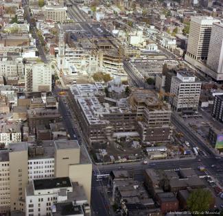 Photographie aérienne couleur du centre-ville avec le chantier de construction de l'université au centre, entourant les églises. 