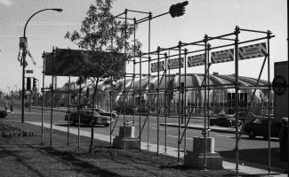 Des échafaudages métalliques et des banderoles annonçant « Corridart dans la rue Sherbrooke » placés sur le trottoir. Derrière, des automobiles circulent. En arrière-plan, le stade olympique de Montréal.