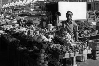 Un commerçant derrière ses étals de légumes au marché montre une aubergine.
