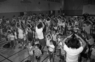 Une foule d'enfants suit les mouvements d'un moniteur dans un gymnase.