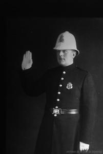Photographie en noir et blanc d’un policier en uniforme. Il a la main droite levée et porte un couvre-chef blanc de forme un peu conique.