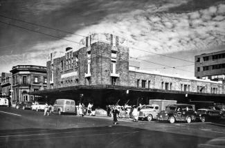 Photo en noir et blanc montrant un édifice abritant un marché vu du coin de la rue. Des voitures et des gens circulent. 