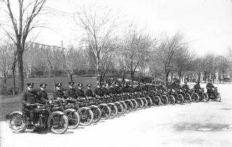 Photographie en noir et blanc d’un groupe de policiers sur leur moto. Ils sont les uns à côté des autres sur le bord de la rue.