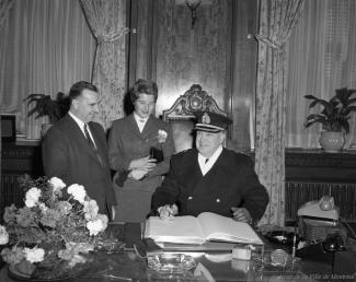 Signature du livre d’or à l’hôtel de ville de Montréal. Le maire Sarto Fournier et une femme sont debout, alors que le chef de police est assis, crayon en main pour signer.