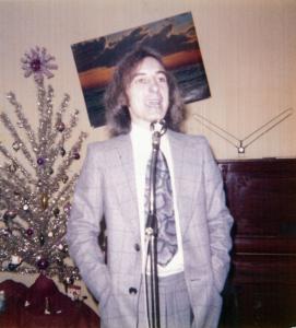 Un homme en veston cravate, cheveux aux épaules, les mains dans les poches, debout devant un micro, a la bouche ouverte. Derrière lui, on voit, sur le mur un cadre croche, le bout d’un piano et un sapin de Noël. 