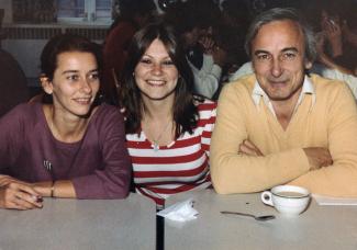 Deux femmes sont attablées avec un homme qui sourit, un café posé devant lui. 