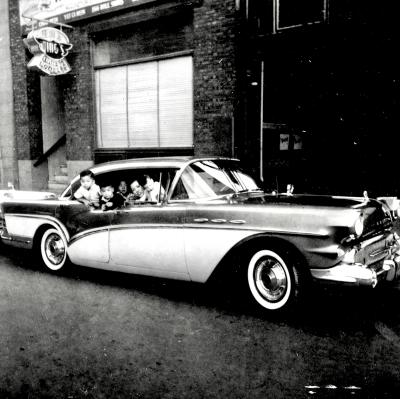 Photo en noir et blanc d'une voiture devant un édifice avec l'enseigne de Wing's Noodle. Une femme et quatre enfants sont dans la voiture aux fenêtres ouvertes.