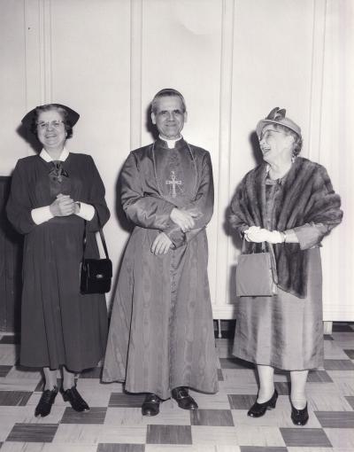 Un homme d’Église est entouré de deux femmes, une habillée d’un costume et l’autre avec fourrure. Les trois ont un sourire, mais la femme de droite sourit plus amplement en regardant les deux autres.