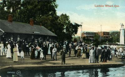 Carte postale colorisée montrant une foule d’hommes, de femmes et d’enfants sur un quai, avec la gare en arrière-plan et des maisons.