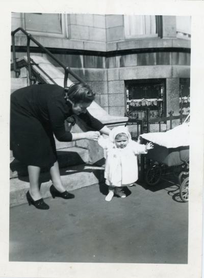 Photo en noir et blanc d’un bébé qui semble faire ses premiers pas sur le trottoir devant la maison avec sa mère qui le tient par la main. Le landau de l’enfant se trouve à droite.  