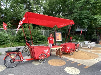 Devant un parc, une femme se tient debout derrière deux vélos dotés de grands coffres et d’une tente de couleur rouge.