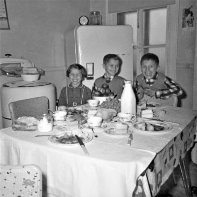 Photo en noir et blanc de trois enfants souriant dans une cuisine avec un réfrigérateur et une machine à laver avec essoreuse. Devant eux, une table bien remplie. 
