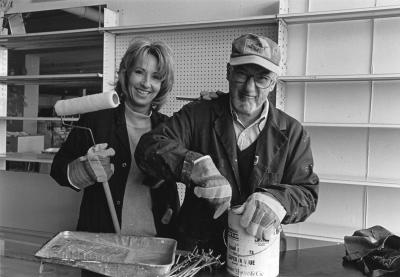 Une femme souriante tient un rouleau de peinture et un homme avec une casquette et des lunettes ouvre un pot de peinture en grimaçant. Derrière eux, des tablettes vides.