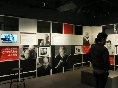 Salle d’exposition montrant deux murs avec des photos en noir et blanc, des panneaux de texte, un écran et deux personnes, debout, qui la visitent. 
