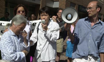 Photo couleur de quelques personnes manifestant. Au centre, une femme tient un micro et un homme à ses côté tient un porte-voix. Une personne âgée pleure à l’avant-plan. 