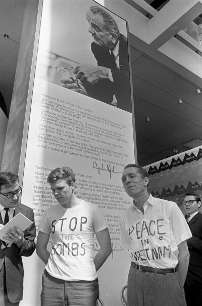 Deux militants anti-guerre du Vietnam portent des chandails avec des messages, l’un écrit « Stop the bombs » et l’autre « Peace in Vietnam ». Un homme à leur gauche leur montre un document. 