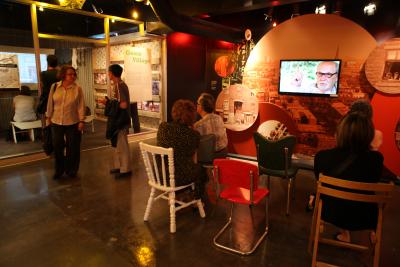 Photo couleur d’une salle d’exposition avec des dispositifs audiovisuels, des panneaux avec photos ou textes et une ambiance tamisée. Sept personnes visitent.
