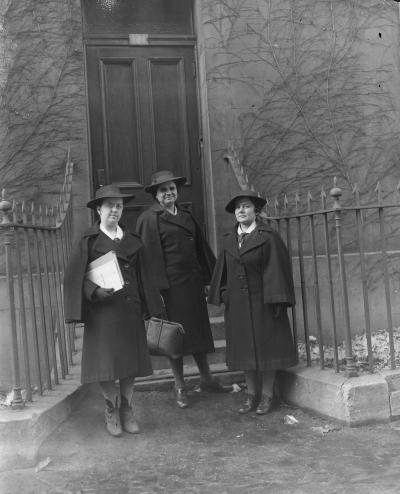 Trois femmes, habillées d’un manteau noir et d’un chapeau, se tiennent devant l’entrée d’une maison. Celle qui est à gauche tient un document et celle du milieu une valise.