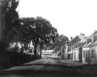 Photo en noir et blanc d’une rue en terre bordée d’arbres d’un côté et de maisons de l’autre. 