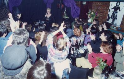 Photo couleur d’un spectacle à l’intérieur d’un bar. Une chanteuse se trouve à l’avant, et devant elle, des femmes assises à des tables applaudissent. 