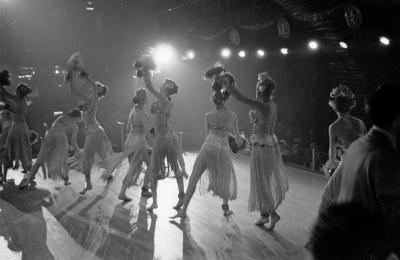 Photographie en noir et blanc d’un groupe de femmes en costume de scène qui dansent. Il y a une vue sur la salle.