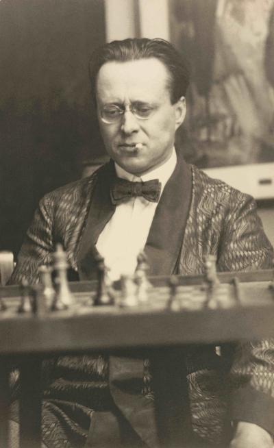 Photographie en noir et blanc d’un homme portant de petites lunettes rondes et un nœud papillon. Une cigarette est pendue au coin droit de sa bouche. Une table est visible devant lui. Sur celle-ci, il y a un jeu d’échecs.