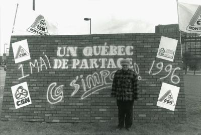 Un homme pose devant une murale où on peut lire le slogan « Un Québec de partage, ça s’impose. 1 mai 1996 ».