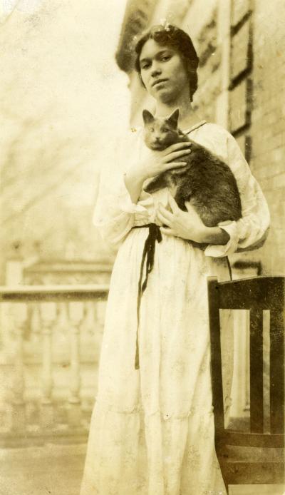Jeune femme, en robe, tenant un chat dans ses bras, sur un balcon d’appartement.