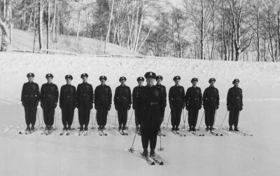 Photographie en noir et blanc sur laquelle on voit des policiers à ski se tenir en ligne droite. L’un d’eux se tient devant la ligne. 