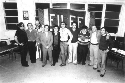 Groupe de neuf hommes posant debout devant des fenêtres d’un local et une banderole sur laquelle est écrit le mot FILEF. 