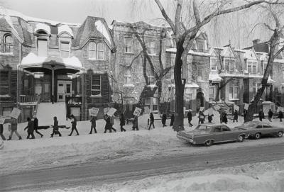 Photo en noir et blanc d’une manifestation dans une rue résidentielle en hiver. Une longue file de personnes se suivent sur le trottoir et tiennent des pancartes.