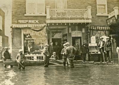Une douzaine d’hommes sont debout devant un commerce. La rue est inondée. Certains ont les pieds dans l’eau, d’autres sont sur une planche ou dans l’entrée d’un commerce. 