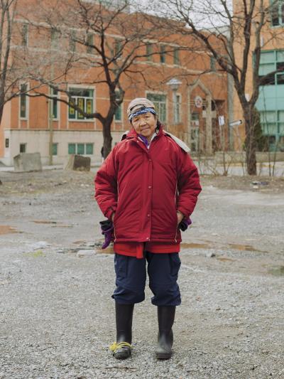 Full length portrait of a woman wearing rubber boots and a cap in a vacant lot in Chinatown. 