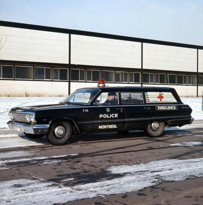 Photographie couleur d’une ambulance noire. Sur la porte du côté conducteur, les mots police et Montréal sont écrits en majuscules. Du même côté, à l’arrière, il y a le mot ambulance. Un gyrophare rouge est visible sur le toit de la voiture.  