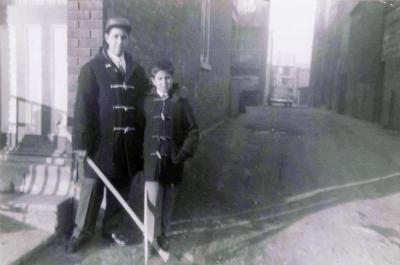 Photo en noir en blanc de 1955 d’un garçon et d’un homme se tenant par la taille dans une ruelle en terre, une voiture se trouve au bout. Ils se tiennent près d’une maison.  