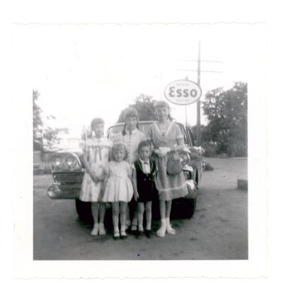 Photographie en noir et blanc montrant un groupe de cinq enfants d’âges variés bien habillés devant une voiture. Il y a un panneau de la compagnie ESSO à l’arrière à droite. 
