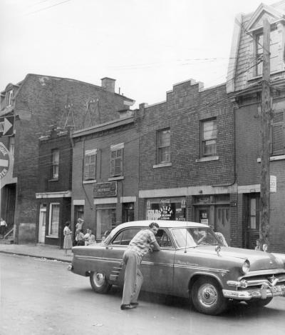Deux jeunes hommes sont accoudés à une voiture stationnée devant une rangée de maisons à deux et trois étages dans la rue de l’Hôtel-de-Ville.