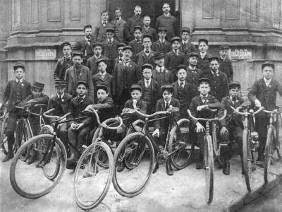 Environ 35 hommes et garçons en rangée posent devant un édifice, avec des vélos en avant-plan.