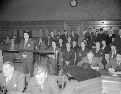 Salle de tribunal remplie d’hommes. À gauche, un homme en complet cravate se tient debout derrière une balustrade. Il a les mains croisées devant lui. 