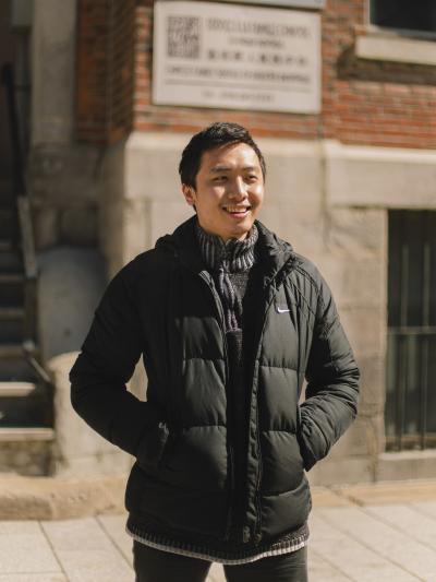A smiling young man, hands in his coat pockets, stands in front of a building in Chinatown. 