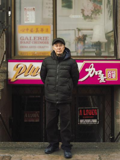 Photo couleur plein pied d’un homme âgé debout devant un édifice commercial du Quartier chinois. 