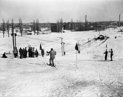 Un skieur portant le numéro 47 est sur le point de descendre une piste. On voit une douzaine de personnes le long de la piste et des édifices en arrière-plan. 