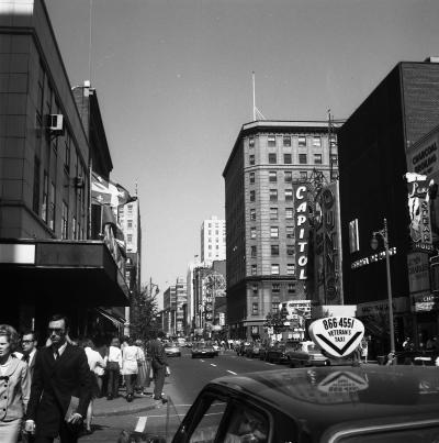 Photo en noir et blanc montrant la rue Sainte-Catherine à la hauteur des rues Peel et Drummond en 1969. On y voit des passants sur le trottoir, les enseignes des magasins, et une voiture de taxi dans le coin inférieur droit de la photo avec un lanternon V