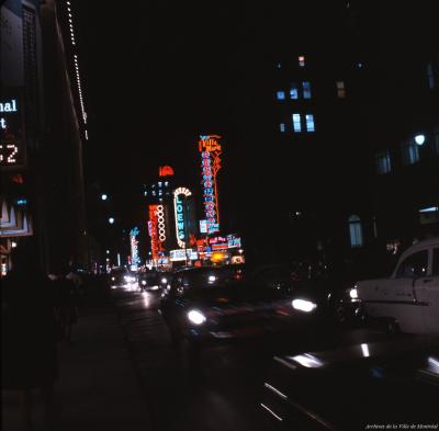 Photo couleur de la rue Sainte-Catherine la nuit avec des enseignes au néon et des voitures au premier plan. 