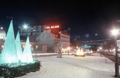 Photo couleur de la place Jacques-Cartier prise de nuit en hiver avec une mince couche de neige. Des décorations en forme de sapins sont placées en groupe de trois le long de la place. L’enseigne de l’hôtel Nelson est illuminée. 